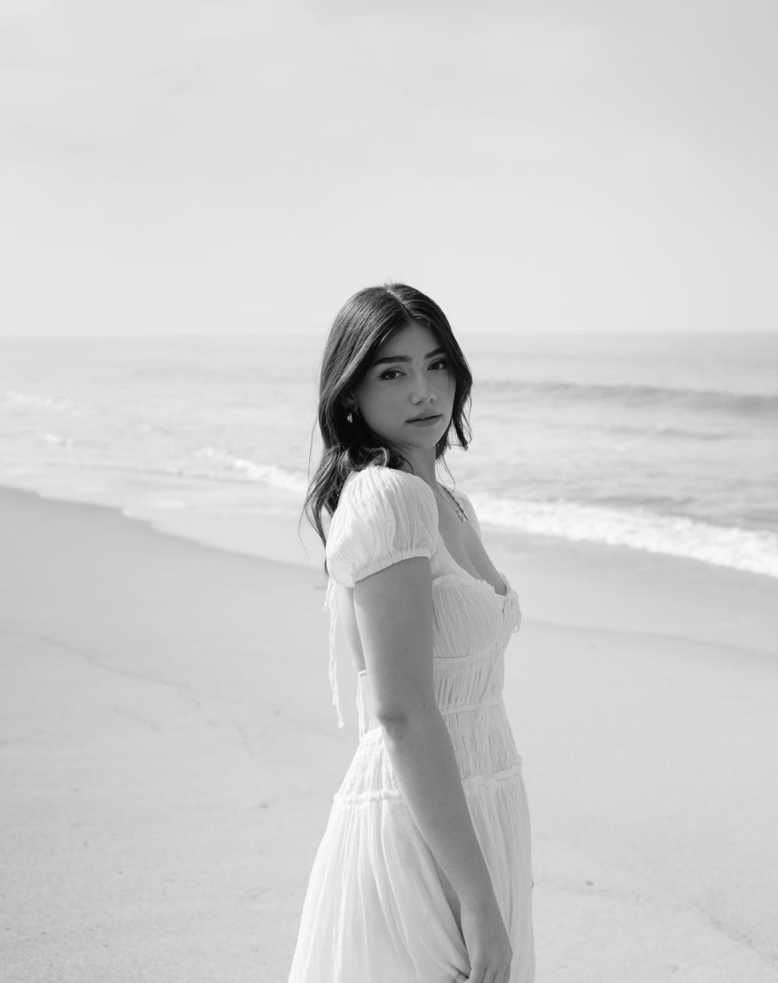 A black and white photo of a young white woman standing on the beach, wearing a white dress and posing in a side profile.